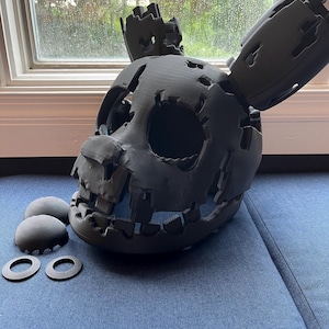 May include: A dark gray, 3D-printed Springtrap mask with visible damage and exposed features. The mask has large eye sockets, a wide mouth, and attached ear pieces. Two matching circular pieces and two dome-shaped pieces are next to the mask.