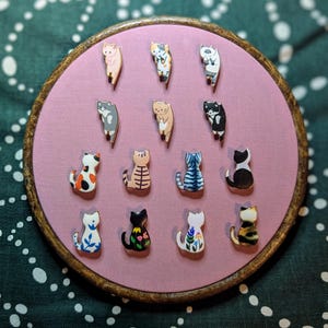 May include: A collection of 14 enamel pins featuring different cat designs. The pins are arranged in a circle on a pink background. The cats are all in different poses and have different expressions. Some of the cats are wearing hats or accessories.
