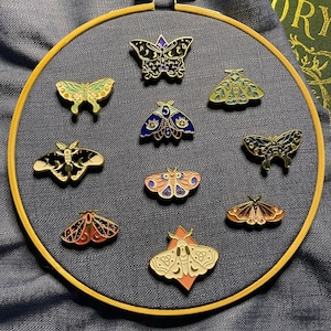 May include: A collection of 10 colourful enamel pins featuring different moth designs. The pins are displayed on a wooden embroidery hoop against a dark blue fabric background.