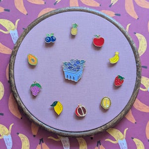 Peut inclure: Un tambour à broder circulaire en bois contenant un tissu violet clair. Dix épinglettes en émail sont disposées en cercle, représentant un ananas, une pomme, une banane, une fraise, une tranche d'agrume, une grenade, un citron, du raisin, une papaye et des myrtilles, avec une épinglette en forme de panier de baies au centre.