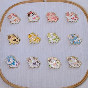 May include: Twelve enamel cow pins with various colours and patterns, displayed on white fabric within a wooden embroidery hoop. Each pin features a different design, creating a diverse collection.