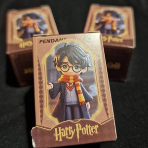 May include: Three small boxes featuring a Harry Potter pendant. The boxes are brown with a cartoon image of Harry Potter in his school uniform. The words "Harry Potter" are printed on the front of the box.