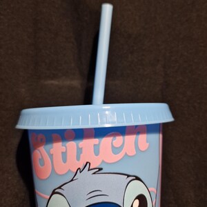 Puede incluir: Un vaso azul claro con tapa y pajita a juego. El vaso presenta una ilustraci&oacute;n de dibujos animados de Stitch de Lilo & Stitch, con la palabra "Stitch" en escritura rosa. La taza est&aacute; dise&ntilde;ada para bebidas fr&iacute;as.