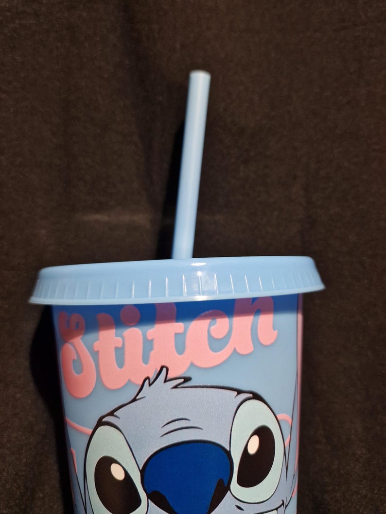 Puede incluir: Un vaso azul claro con tapa y pajita a juego. El vaso presenta una ilustraci&oacute;n de dibujos animados de Stitch de Lilo & Stitch, con la palabra "Stitch" en escritura rosa. La taza est&aacute; dise&ntilde;ada para bebidas fr&iacute;as.