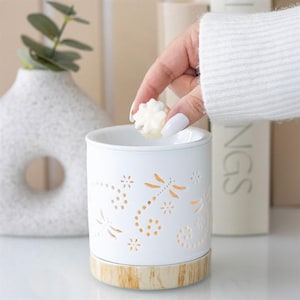 May include: White ceramic wax warmer with dragonfly cutouts and a wooden base. A hand is placing a white wax melt into the warmer. The warmer is cylindrical and has a decorative design. The background includes a vase and books.