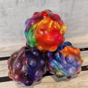 May include: Three spherical sensory toys with a dimpled, bubble-like surface texture. The top sphere features a vibrant swirl of red, orange, yellow, and green. The bottom left sphere is deep purple, and the bottom right sphere has a blue and orange swirl.
