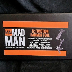 May include: A black box with an orange stripe, displaying the text "MM MAD MAN" and "12 FUNCTION HAMMER TOOL." The box lists features including a saw blade, bottle opener, wire cutters, and pliers. The text "GIFTS GADGETS & GEAR FOR GUYS" is also visible.