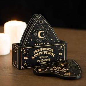 May include: A black wooden box containing triangular coasters with gold celestial designs, including stars, moons, and an eye. The box features the alphabet, numbers 0-9, the words YES, NO, and GOODBYE, and a planchette-shaped coaster rests in front.