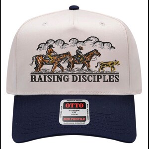 Puede incluir: Gorra de b&eacute;isbol con corona color crema y visera azul marino. La gorra presenta una ilustraci&oacute;n de vaqueros a caballo y el texto "RAISING DISCIPLES". La gorra tiene un ajuste cl&aacute;sico y est&aacute; hecha de tela de primera calidad.