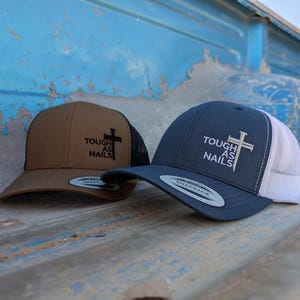 May include: Two baseball caps with a black cross and the text "Tough as Nails" embroidered on the front. One cap is brown with a black mesh back, and the other is navy blue with a white mesh back.