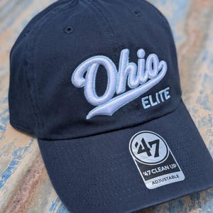 May include: A navy blue baseball cap featuring the word Ohio embroidered in light blue script with a white outline, and the word ELITE in white block letters. A black and white sticker on the brim displays the number 47 and the text 47 CLEAN UP ADJUSTABLE.