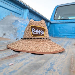 May include: A straw hat with a wide brim and a brown and white patch that reads "Briggs". The hat has a black and white rope detail around the crown and a brown trim. The hat is resting on the bed of a blue truck.