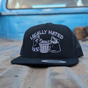 May include: Black trucker hat with a flat brim. The hat features the text "LOCALLY HATED" in white, along with a raccoon and bear graphic next to a trash can. The hat has a mesh back and a snapback closure.