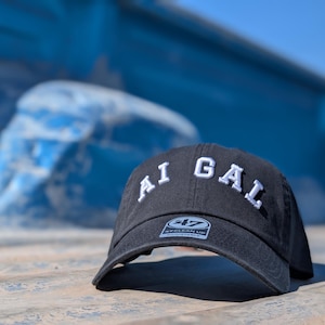Könnte beinhalten: Schwarze Baseballkappe mit dem weißen Text "AI GAL", mit gebogenem Schirm und einem 47 Clean Up-Etikett. Die Kappe liegt auf einer strukturierten Oberfläche mit verschwommenem blauem Hintergrund.