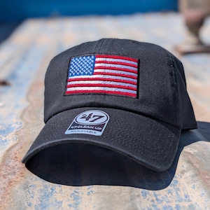 May include: A black baseball cap featuring an embroidered rectangular American flag patch on the front panel. The cap has a curved brim with a circular sticker that reads 47 CLEAN UP ADJUSTABLE in white text on a dark background.