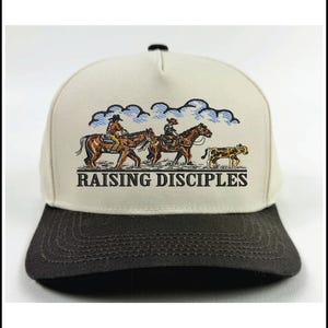 Puede incluir: Gorra de b&eacute;isbol beige y gris oscuro con una ilustraci&oacute;n de dos jinetes a caballo y un leopardo, con nubes azules de fondo. El texto "RAISING DISCIPLES" est&aacute; impreso debajo.