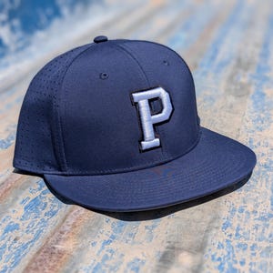 May include: Navy blue baseball cap with a flat brim. The cap features a white and gray embroidered letter "P" on the front. The cap has a perforated design on the side panels for breathability.