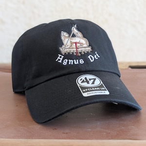 Może przedstawiać: Czarna czapka bejsbolówka z haftowanym wizerunkiem baranka i napisem "Agnus Dei" z przodu. Czapka ma białą etykietę "47 Clean Up Adjustable" z boku.
