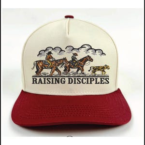 Puede incluir: Gorra de b&eacute;isbol con corona color crema y visera granate. La gorra presenta una ilustraci&oacute;n de vaqueros a caballo con el texto "RAISING DISCIPLES" en negro.
