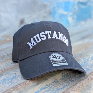 Op de afbeelding: Een antracietgrijze honkbalpet met het woord MUSTANGS in witte gebogen letters op de voorkant geborduurd. De pet heeft een ronde zwart-witte sticker op de klep met de tekst 47 CLEAN UP ADJUSTABLE.