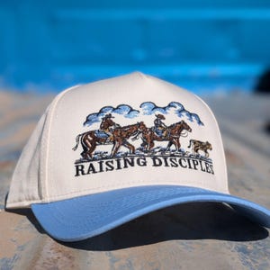 Puede incluir: Gorra de b&eacute;isbol color crema con visera azul claro. La gorra presenta un dise&ntilde;o bordado de vaqueros a caballo bajo un cielo nublado, con las palabras "RAISING DISCIPLES" en negro.
