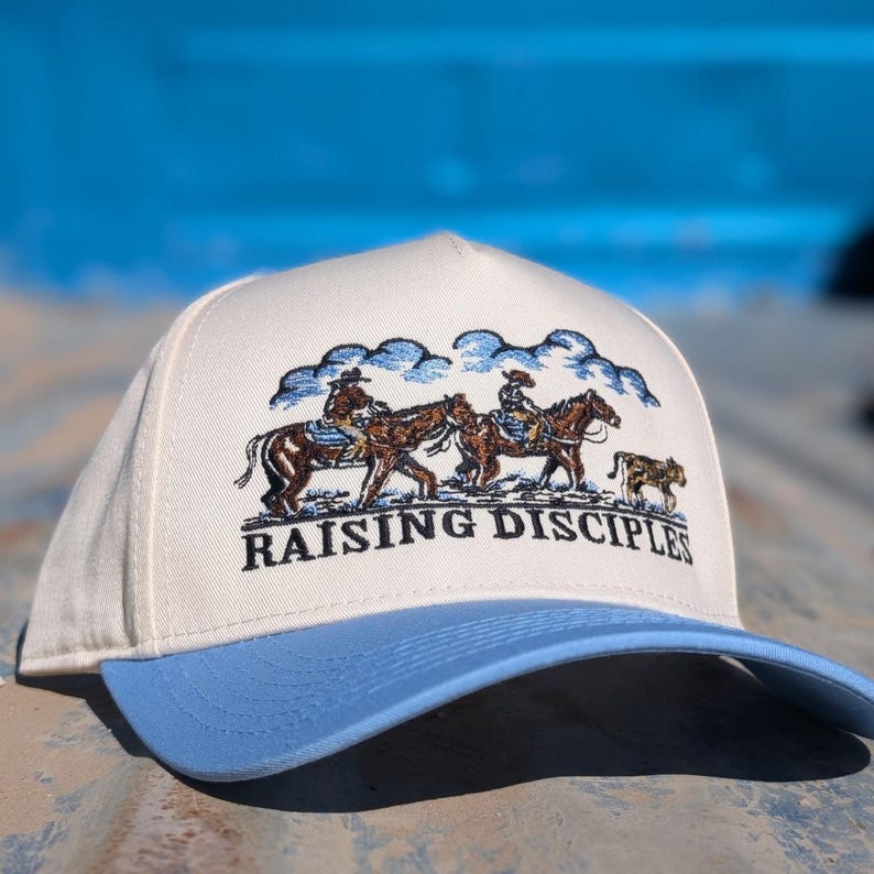 Puede incluir: Gorra de b&eacute;isbol color crema con visera azul claro. La gorra presenta un dise&ntilde;o bordado de vaqueros a caballo bajo un cielo nublado, con las palabras "RAISING DISCIPLES" en negro.