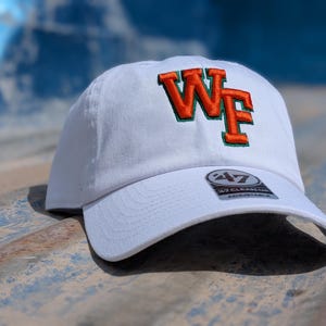 Peut inclure: Une casquette de baseball blanche avec les lettres brodées WF en orange et un contour vert sur le devant. La casquette comporte un autocollant circulaire sur la visière avec le texte 47 CLEAN UP ADJUSTABLE, indiquant la marque et le style.