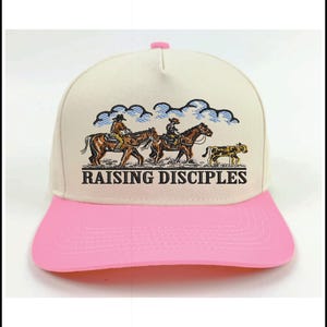 Puede incluir: Gorra de b&eacute;isbol color crema con visera y bot&oacute;n superior rosas. La gorra presenta una ilustraci&oacute;n de jinetes a caballo con el texto "RAISING DISCIPLES" en negro.