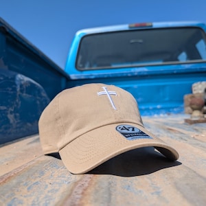 Może przedstawiać: Beżowa czapka z daszkiem z białym krzyżem wyhaftowanym z przodu. Czapka ma zakrzywiony daszek i logo z boku. Leży na zwietrzałej, zardzewiałej powierzchni, z niebieską skrzynią ładunkową w tle.