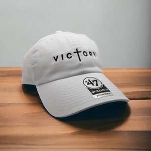 以下が含まれることがあります： 黒色の刺繍で「VICTORY」と刺繍された白い野球帽。Oの上に十字架があります。帽子には、黒と白の「47 Clean Up Adjustable」のラベルが側面に付いています。