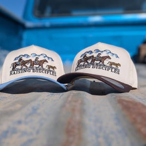 Puede incluir: Dos gorras de b&eacute;isbol beige con visera curva. Cada gorra presenta un dise&ntilde;o bordado de jinetes a caballo y el texto "RAISING DISCIPLES". Una gorra tiene una visera azul, la otra una visera marr&oacute;n.