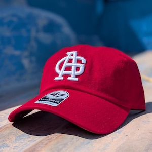 Puede incluir: Gorra de béisbol roja con un logotipo blanco bordado. La gorra tiene una visera curva y una etiqueta que dice "47 Clean Up Adjustable". El sombrero está hecho de un material suave.