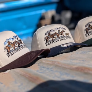 Puede incluir: Tres gorras de camionero beige con diseños bordados de vaqueros y caballos. Cada gorra tiene una visera de diferente color: marrón, azul marino y verde oscuro. El texto "RAISING DISCIPLES" está bordado en la parte delantera de cada gorra.