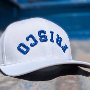 May include: A white baseball cap with the word "FRISCO" embroidered in blue, arched across the front. The cap has a curved brim and a structured fit, suitable for casual wear.