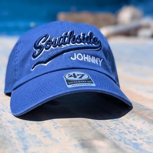 May include: A blue baseball cap with the word "Southside" embroidered in white script above the name "Johnny". The cap has a curved brim and an adjustable strap. The "47 Clean Up" logo is visible on the front.