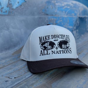 May include: White baseball cap with a brown brim. The front of the cap has black embroidery that reads "MAKE DISCIPLES OF ALL NATIONS" with two globes and a cross.
