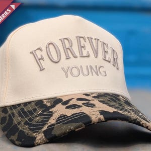 May include: A cream-colored baseball cap featuring the embroidered text FOREVER YOUNG in a curved arch. The cap has a structured crown and a curved brim with a black, brown, and tan leopard print pattern. The design includes a red corner banner.