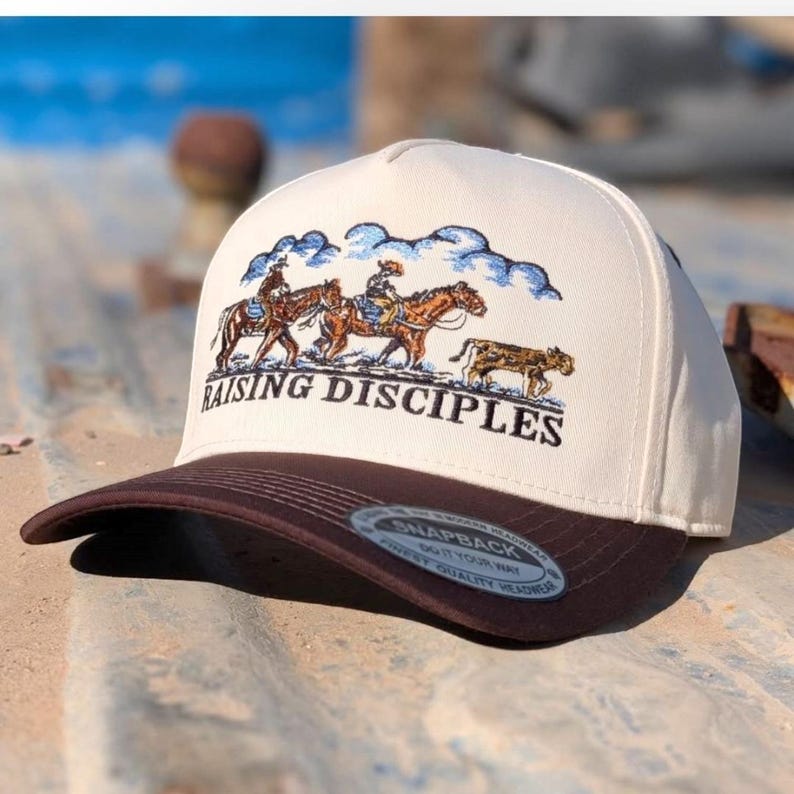 Puede incluir: Gorra snapback color crema con visera marr&oacute;n. La gorra presenta un dise&ntilde;o bordado de tres jinetes a caballo con el texto "RAISING DISCIPLES" en negro. El fondo incluye nubes azules.