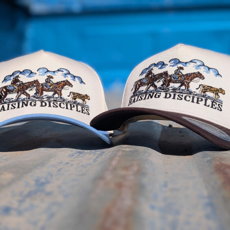 Puede incluir: Dos gorras de b&eacute;isbol con corona color crema y visera curva. Las gorras presentan un dise&ntilde;o bordado de vaqueros a caballo y el texto "RAISING DISCIPLES". Una gorra tiene la visera marr&oacute;n, la otra azul claro.