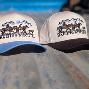 Puede incluir: Dos gorras de b&eacute;isbol con corona color crema y visera curva. Una tiene visera azul claro, la otra marr&oacute;n oscuro. Ambas gorras presentan un dise&ntilde;o bordado de vaqueros a caballo con el texto "RAISING DISCIPLES".