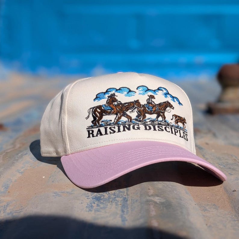 Puede incluir: Gorra de b&eacute;isbol color crema con visera lavanda. La gorra presenta un dise&ntilde;o bordado de jinetes a caballo con el texto "RAISING DISCIPLES" en negro.