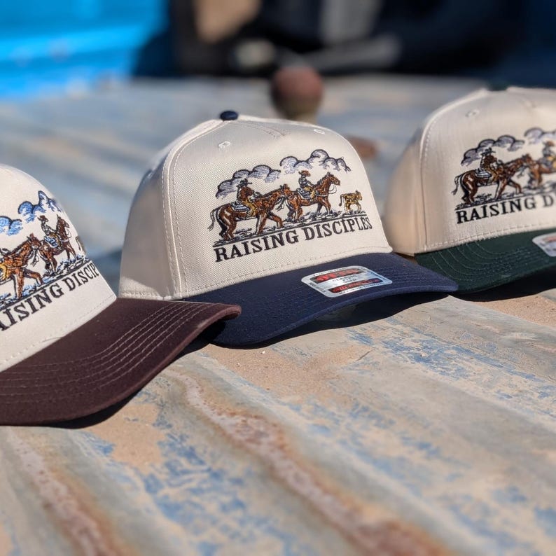 Puede incluir: Tres gorras de b&eacute;isbol con corona color crema y visera oscura. Cada gorra presenta un dise&ntilde;o bordado de jinetes a caballo y el texto "RAISING DISCIPLES". Las viseras son marrones, azul marino y verde oscuro.