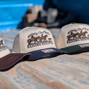 Puede incluir: Tres gorras de b&eacute;isbol con corona crema, visera azul oscuro y un gr&aacute;fico de jinetes a caballo. El texto "RAISING DISCIPLES" est&aacute; debajo del gr&aacute;fico. Una gorra tiene visera marr&oacute;n, y otra visera verde oscuro.