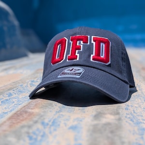 May include: A dark navy blue baseball cap featuring the letters OFD embroidered in bold red with a white outline on the front panel. The cap includes a circular 47 brand sticker on the brim with the text 47 CLEAN UP ADJUSTABLE visible.