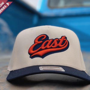 Peut inclure: Casquette de baseball beige et bleu marine avec le mot "East" en lettres orange et bleu marine. La casquette a une visière plate et une couronne structurée. Le texte "Retro Drop Shadow Design Series" est dans le coin supérieur gauche.