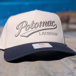May include: A cream-colored, flat-brimmed cap with a navy blue brim. The word "Potomac" is embroidered in a cursive font above the word "Lacrosse". The cap has a structured fit and a tag on the brim.