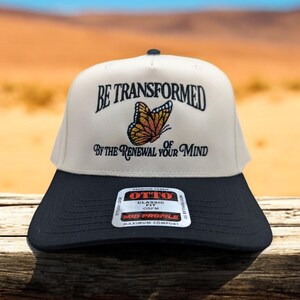 Peut inclure: Une casquette de baseball beige et noire avec le texte "BE TRANSFORMED BY THE RENEWAL OF YOUR MIND" et un graphique de papillon. La casquette a une visière noire et une couronne blanche. La casquette est fabriquée par OTTO et a une coupe classique et un profil moyen.