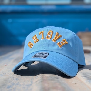 May include: A light blue baseball cap featuring the word EAGLES embroidered in gold thread with a white outline across the front. The cap includes a 47 Clean Up adjustable sticker on the brim and has a structured crown with a curved visor.