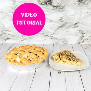 May include: Two apple pies, one whole and one sliced, on a white wooden surface. The whole pie is in a silver pan. The sliced pie is on a white plate. A pink circle with the text "VIDEO TUTORIAL" is in the top left corner of the image.