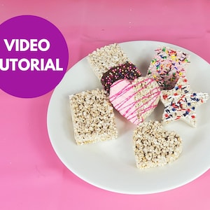 May include: A white plate with six rice crispy treats shaped as a heart, star, and squares. The treats are decorated with pink, red, blue, and white sprinkles and chocolate.